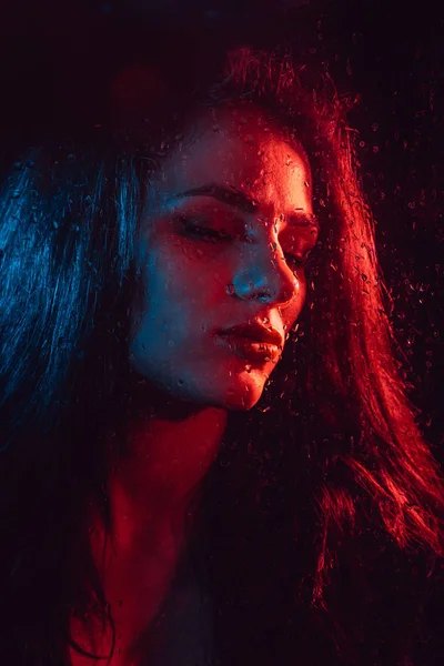 Sensual portrait of sad girl through glass with raindrops with red blue lighting — Stock Photo, Image