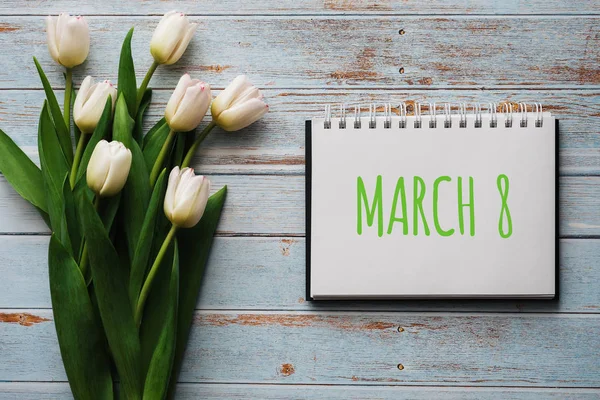 Ramo de flores blancas de tulipanes en el fondo de las tablas azules con un cuaderno con letras 8 de marzo — Foto de Stock
