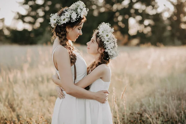 Sorelle felici in abiti bianchi con ghirlande floreali e trecce in stile boho in estate in un campo — Foto Stock