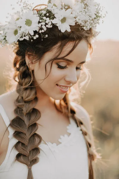 Retrato de menina sorridente em vestido branco com coroa floral e tranças no verão ao pôr do sol no campo — Fotografia de Stock