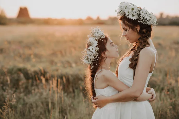 Mãe e filha abraçando juntos em vestidos brancos com tranças e coroas florais no estilo boho no verão — Fotografia de Stock