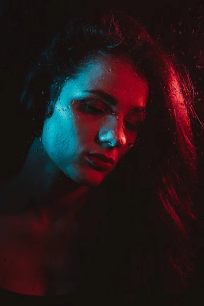 Zmysłowy portret pięknej dziewczyny za szybą z krople deszczu z czerwony niebieski oświetlenie — Zdjęcie stockowe