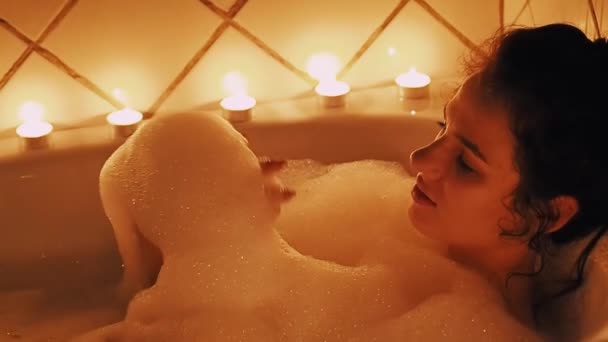 Chica joven se relaja juguetonamente en el baño y sopla de la espuma — Vídeo de stock