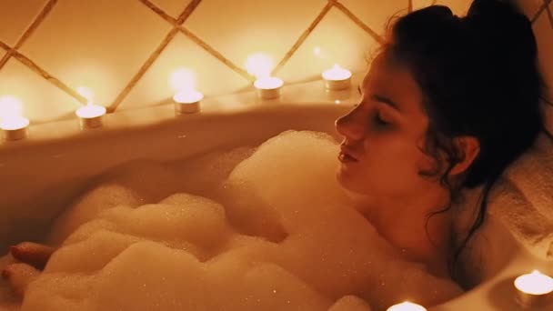 Young brunette girl enjoys and relaxes in a bubble bath with candles and smiles happily — 비디오