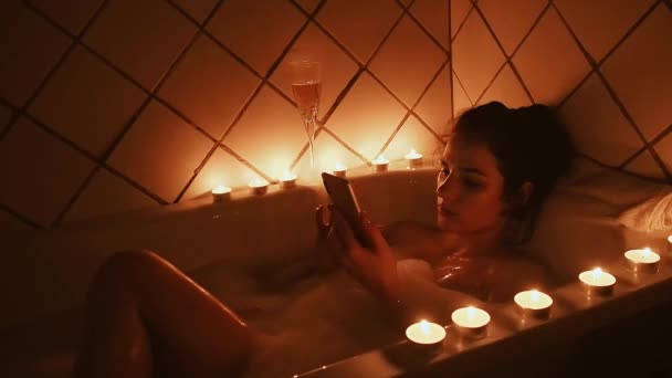 Gelooid sexy jong meisje ligt in een schuimbad met een smartphone en ziet er met een glimlach op het scherm van de telefoon flipping door de tape — Stockvideo