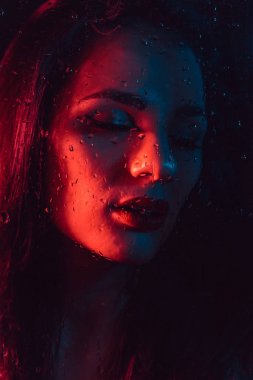 Yağmur damlaları ile cam arkasında kırmızı mavi aydınlatmalı üzgün genç kız portresi