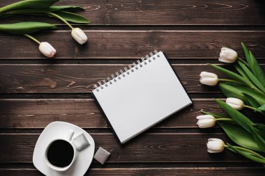 Bir fincan kahve ve boş bir not defteri ile ahşap bir masa üzerinde beyaz laleler