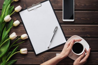 Boş bir kağıt tablet, akıllı telefon ve kadınların elinde bir fincan kahveyle ahşap bir masada beyaz laleler.