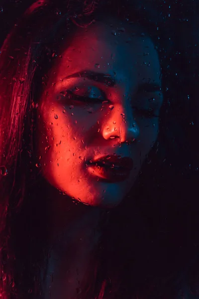 Portret smutny młoda dziewczyna z czerwony niebieski oświetlenie za szybą z deszczu — Zdjęcie stockowe
