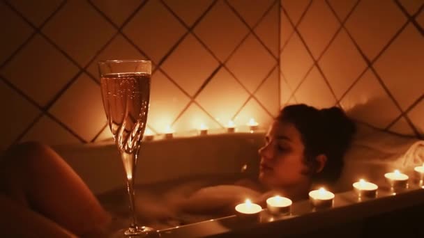 Glas Champagner mit Kerzen auf dem Hintergrund eines unscharfen entspannten jungen Mädchens im Badezimmer — Stockvideo
