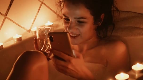 Молодая девушка принимает ванну с пеной со смартфоном, смеется радостно касаясь экрана — стоковое видео
