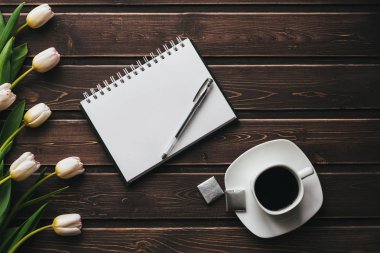 Bir fincan kahve ve boş bir not defteri ile ahşap bir masa üzerinde beyaz laleler