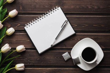 Bir fincan kahve ve boş bir not defteri ile ahşap bir masa üzerinde beyaz laleler