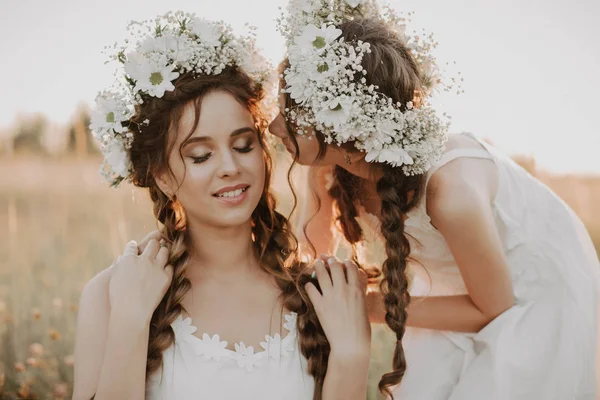 Gelukkige moeder en dochter glimlachen en knuffelen in het veld in de zomer in witte jurken met vlechten en bloemenkransen — Stockfoto