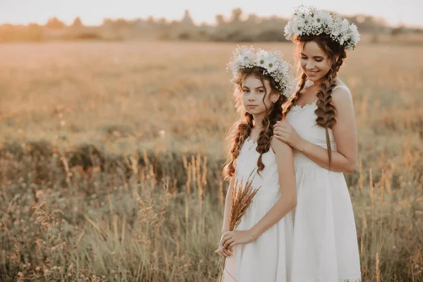 Mãe e filha juntas em vestidos brancos com tranças e coroas florais em estilo boho no campo de verão ao pôr do sol — Fotografia de Stock