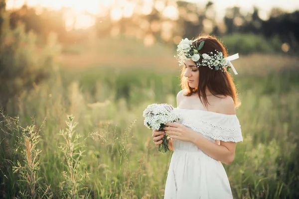 Vacker ung flicka med en bukett blommor och en krans i en vit klänning — Stockfoto