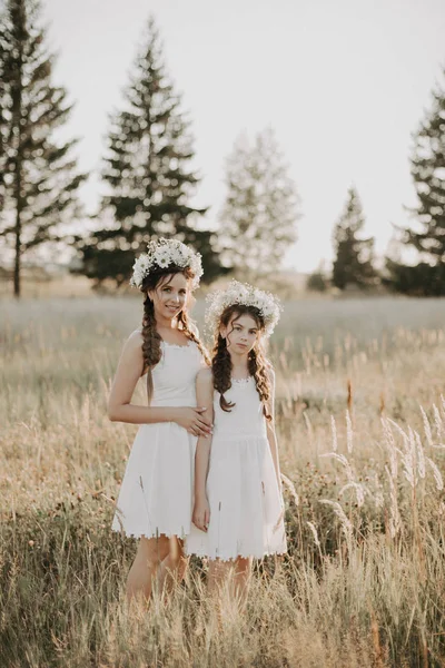 Feliz mamá e hija en vestidos blancos con coronas florales y trenzas de estilo boho en verano en un campo — Foto de Stock