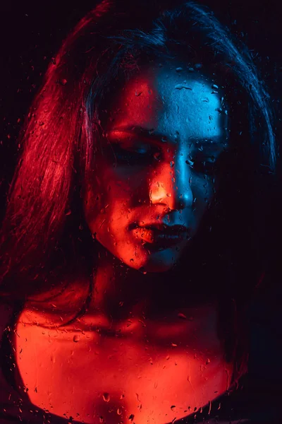 Portret zmysłowy smutną dziewczynę przez szkło z krople deszczu z czerwony niebieski oświetlenie — Zdjęcie stockowe