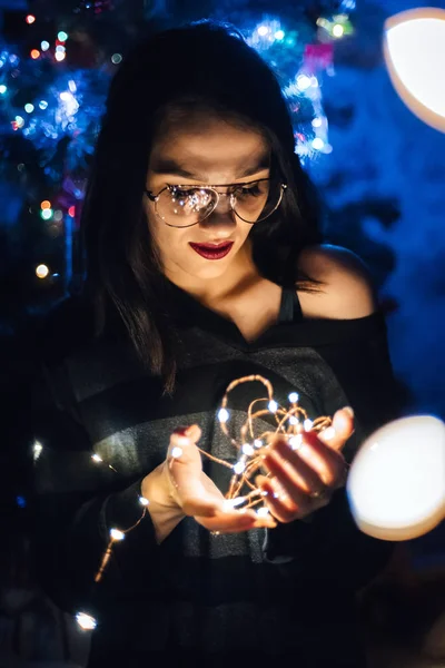 Κορίτσι με γυαλιά που κρατούν ένα στεφάνι με φώτα τη νύχτα στο σπίτι — Φωτογραφία Αρχείου