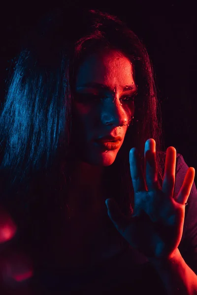 Sensual portrait of sad girl through glass with raindrops with red blue lighting — Stock Photo, Image