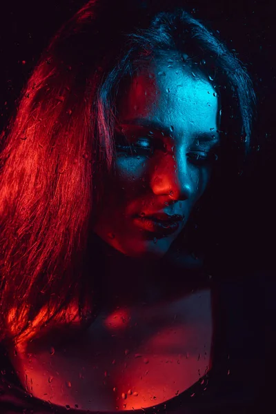 Portret smutny młoda dziewczyna z czerwony niebieski oświetlenie za szybą z deszczu — Zdjęcie stockowe