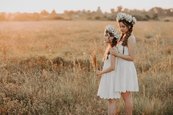 Mãe e filha juntas em vestidos brancos com tranças e coroas florais em estilo boho no campo de verão ao pôr do sol — Fotografia de Stock