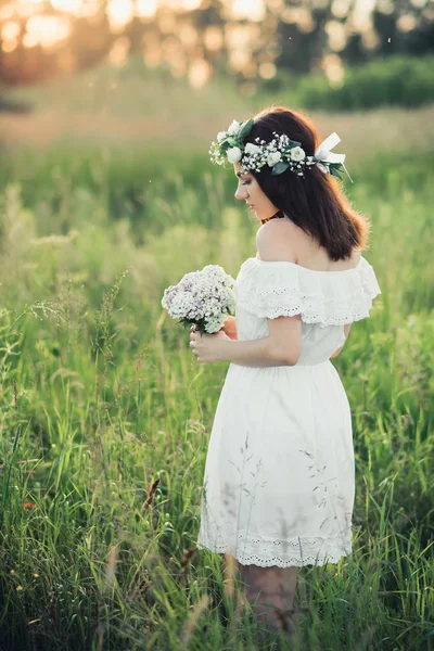 Bella ragazza con un mazzo di fiori e una ghirlanda in un vestito bianco — Foto Stock