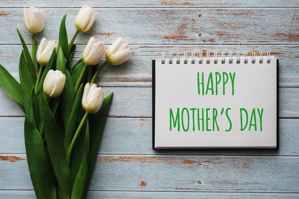 Bukett vita blommor tulpaner på bakgrunden av blå styrelser med en anteckningsbok med bokstäver lycklig mors dag — Stockfoto