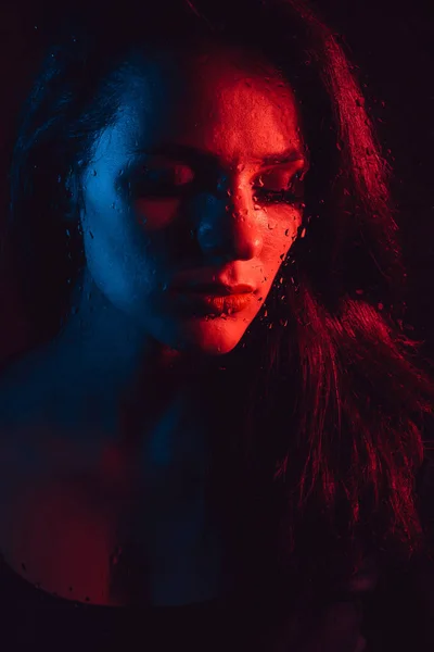 Sensuele portret van verdrietig meisje door glas met regendruppels met rode blauwe verlichting — Stockfoto
