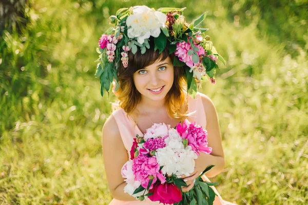 Portret van een vrolijk glimlachend meisje met een krans van bloemen en een boeket van pioenrozen — Stockfoto