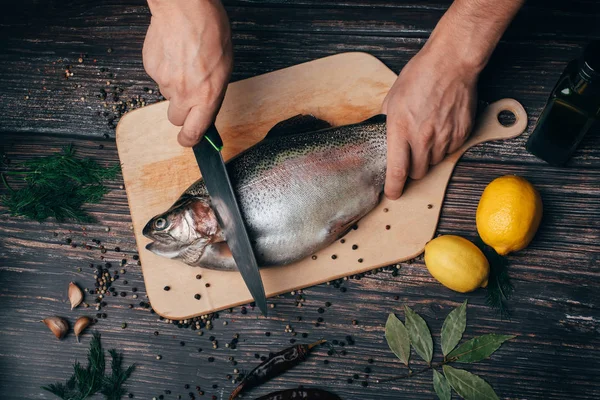 Manos del chef con un cuchillo cortando pescado en una mesa de madera en la cocina. Trucha fresca —  Fotos de Stock