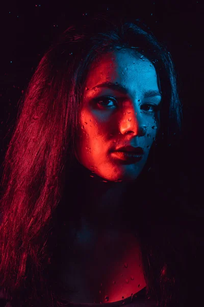 Sensual portrait of beautiful girl behind glass with raindrops with red blue lighting — Stock Photo, Image