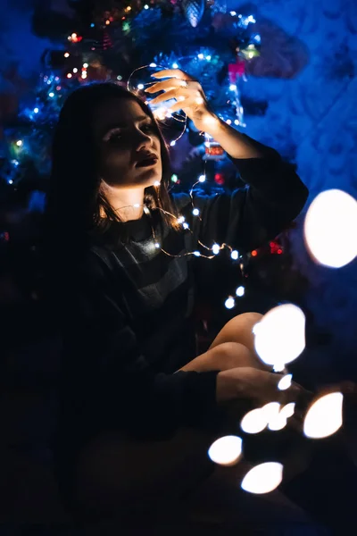 Красивая девушка сидит на фоне праздничной елки с гирляндой — стоковое фото