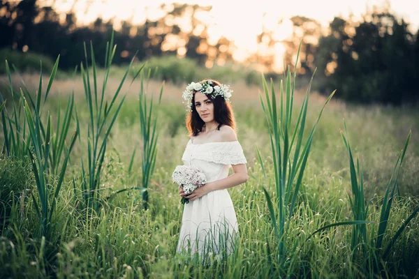 Vacker ung flicka med en bukett blommor och en krans i en vit klänning — Stockfoto
