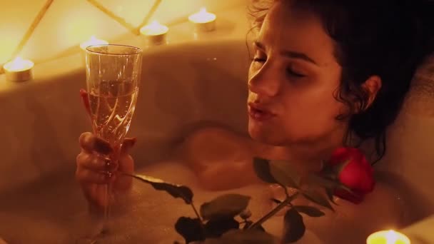 Ευτυχισμένο όμορφο νεαρό κορίτσι που βρίσκεται στο μπάνιο με αφρό με ένα τριαντάφυλλο στο χέρι της και πίνοντας σαμπάνια — Αρχείο Βίντεο
