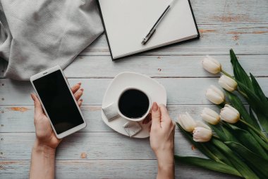 bir Fincan kahve ve kadın elinde bir akıllı telefon ve ahşap bir masa üzerinde boş bir dizüstü bilgisayar ile beyaz lale buketi