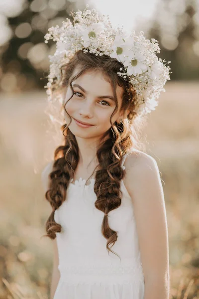 Portrait of a smiling young girl in a white dress and a floral wreath on her hair with braids in the summer field — Stock Photo, Image