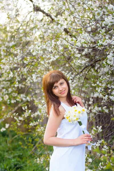 Bella ragazza con un mazzo di narcisi in un vestito bianco su uno sfondo di fiori di ciliegio — Foto Stock