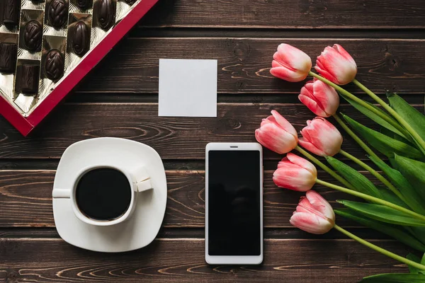 Strauß rosa Tulpenblüten mit einer Tasse Kaffee und einer Schachtel Pralinen und einem Smartphone — Stockfoto