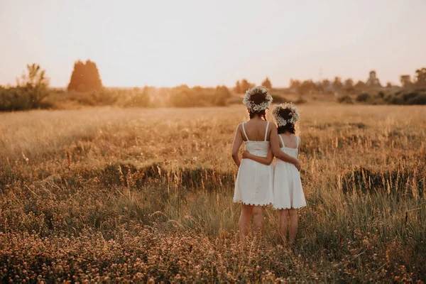 Mother and daughter stand back in the summer at sunset in the field in white dresses and floral wreaths