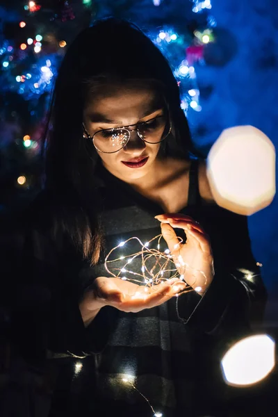 Ragazza con gli occhiali che tiene una ghirlanda con le luci di notte a casa — Foto Stock