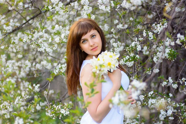 Beautiful young girl with a bouquet of daffodils in a white dress on a background of cherry blossoms — Stock Photo, Image