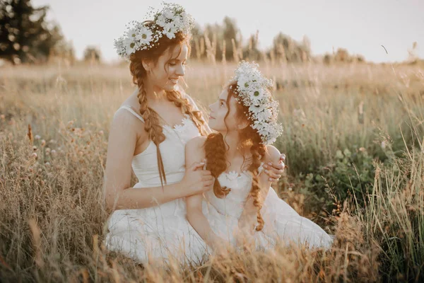 Gelukkige moeder en dochter glimlachen en knuffelen in het veld in de zomer in witte jurken met vlechten en bloemenkransen — Stockfoto