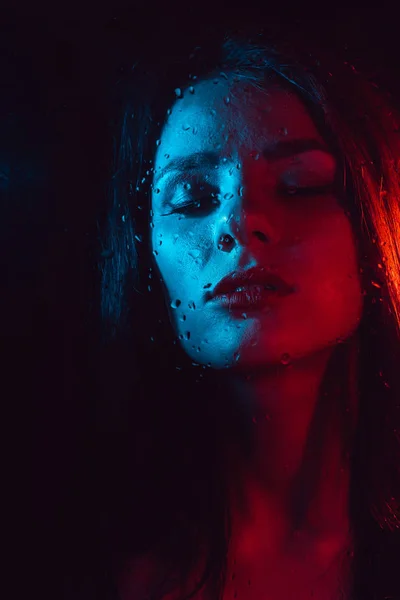 Чувственный портрет красивой девушки за стеклом с капельками дождя с красно-синим освещением — стоковое фото