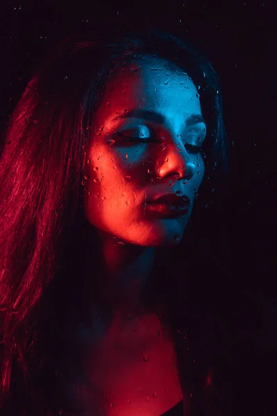 Portrait of sad young girl with red blue lighting behind glass with raindrops — Stock Photo, Image
