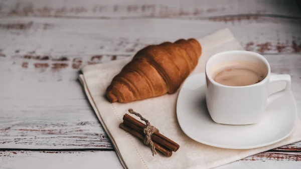 Cup of coffee and croissant with cinnamon sticks on napkin on a wooden table