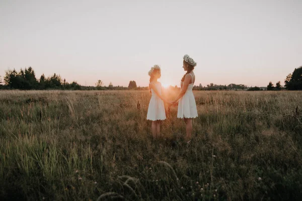 Мать и дочь вместе в белых платьях с косичками и цветочными венками в стиле бохо на летнем поле на закате — стоковое фото