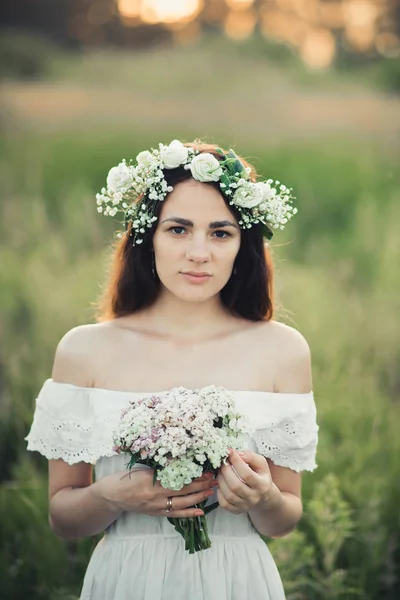 Portret z atrakcyjną dziewczyną w białej sukni z bukietem kwiatów i wieniec w lecie — Zdjęcie stockowe