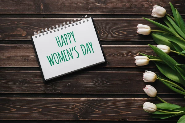 Flores blancas tulipanes sobre tablas de madera marrón. Tarjeta de felicitación con letras Feliz Día de la Mujer — Foto de Stock