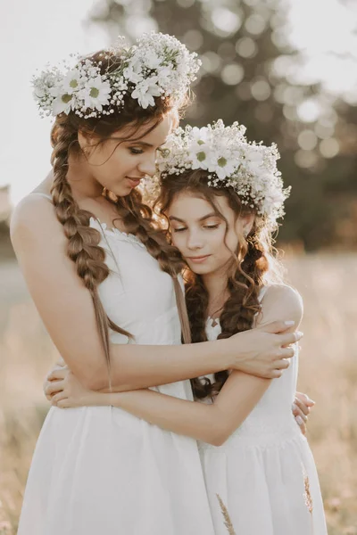 Hermanas felices en vestidos blancos con coronas florales y trenzas de estilo boho en verano en un campo — Foto de Stock